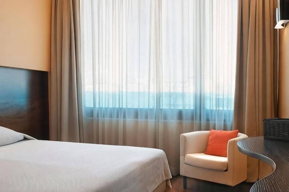 A hotel room with a bed, a chair, a table, and a view of the sea through sheer curtains.