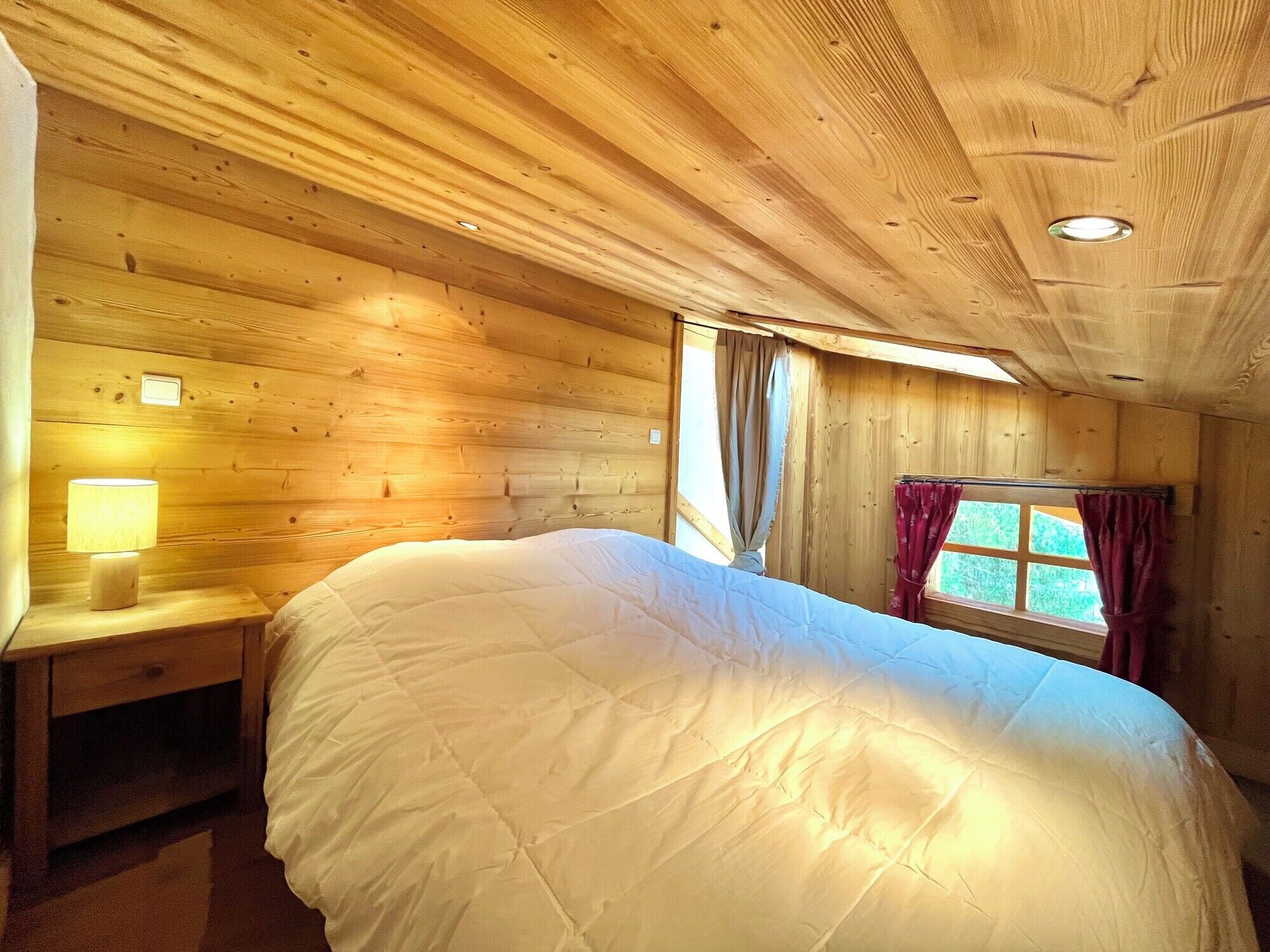 Habitación de cabaña de madera con cama, mesita de noche, ventana con cortinas y lámpara fijada a la pared.