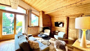 Interior acolhedor de uma cabana de toras, com um sofá, poltronas, uma TV e uma lareira.