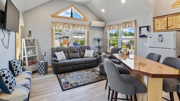 A modern living room with a sofa, chairs, a coffee table, and a TV. There is a large window with curtains, a ceiling fan, and a kitchen area with cabinets. - 3 Usen Lane near all the fun (Old Orchard Beach)