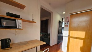 Una cocina compacta con microondas, hervidor de agua y una pequeña mesa con una silla. Se ve un sofá al fondo, y una puerta con un cartel está a la derecha.