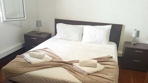 Una cama bien hecha con cubrecama acolchada, dos almohadas y una toalla doblada a cada lado. Hay dos mesitas de noche con lámparas.