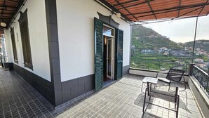 Un balcón con vistas a un paisaje montañoso, una mesa con sillas y una puerta con contraventanas verdes.