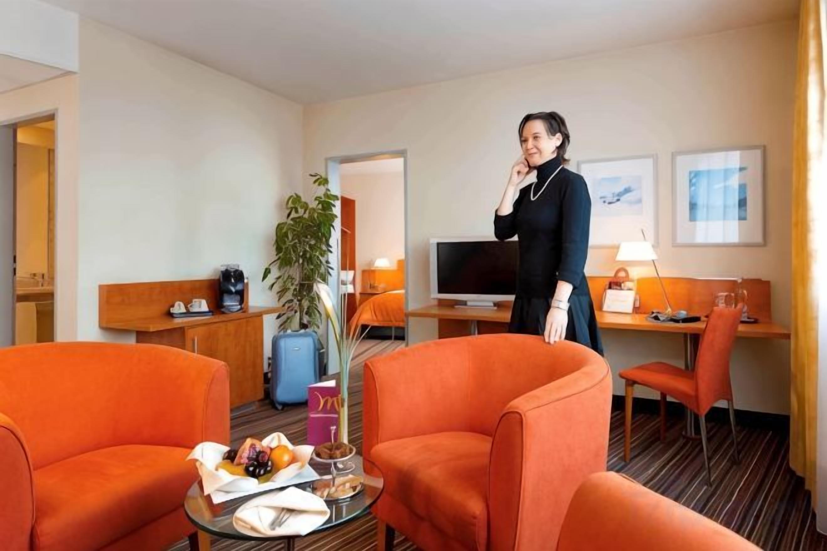 A hotel room with two orange armchairs, a small table with a fruit bowl, a desk with a computer, and a television.