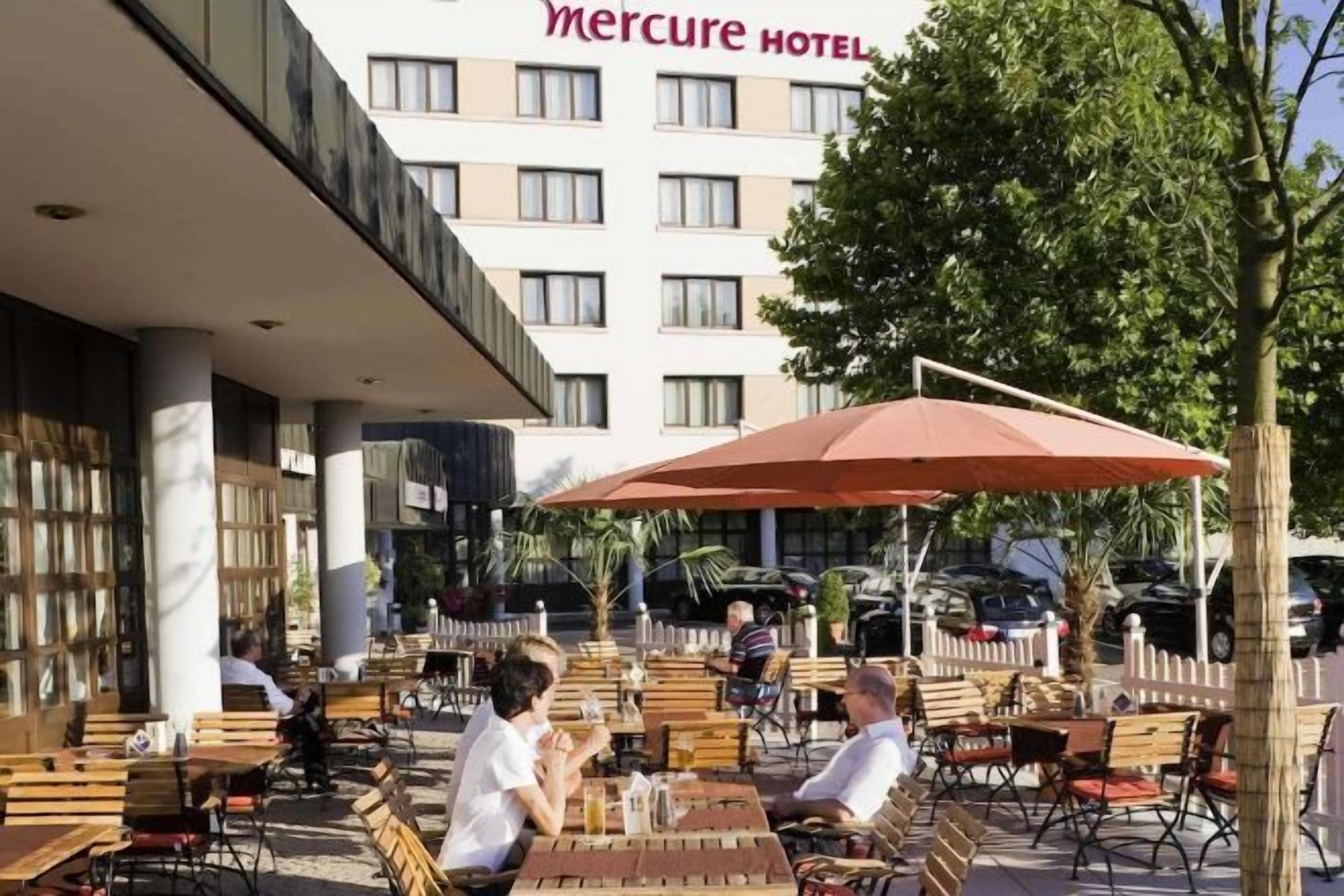 Un área de estar al aire libre con mesas, sillas y sombrillas, y un edificio con la leyenda 'Mercure HOTEL' al fondo.