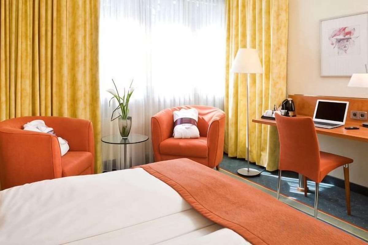 A hotel room with a bed, two armchairs, a desk with a laptop, a lamp, and a window with curtains.