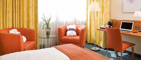 Quarto de hotel com cama, duas poltronas, escrivaninha com laptop, abajur e janela com cortinas.