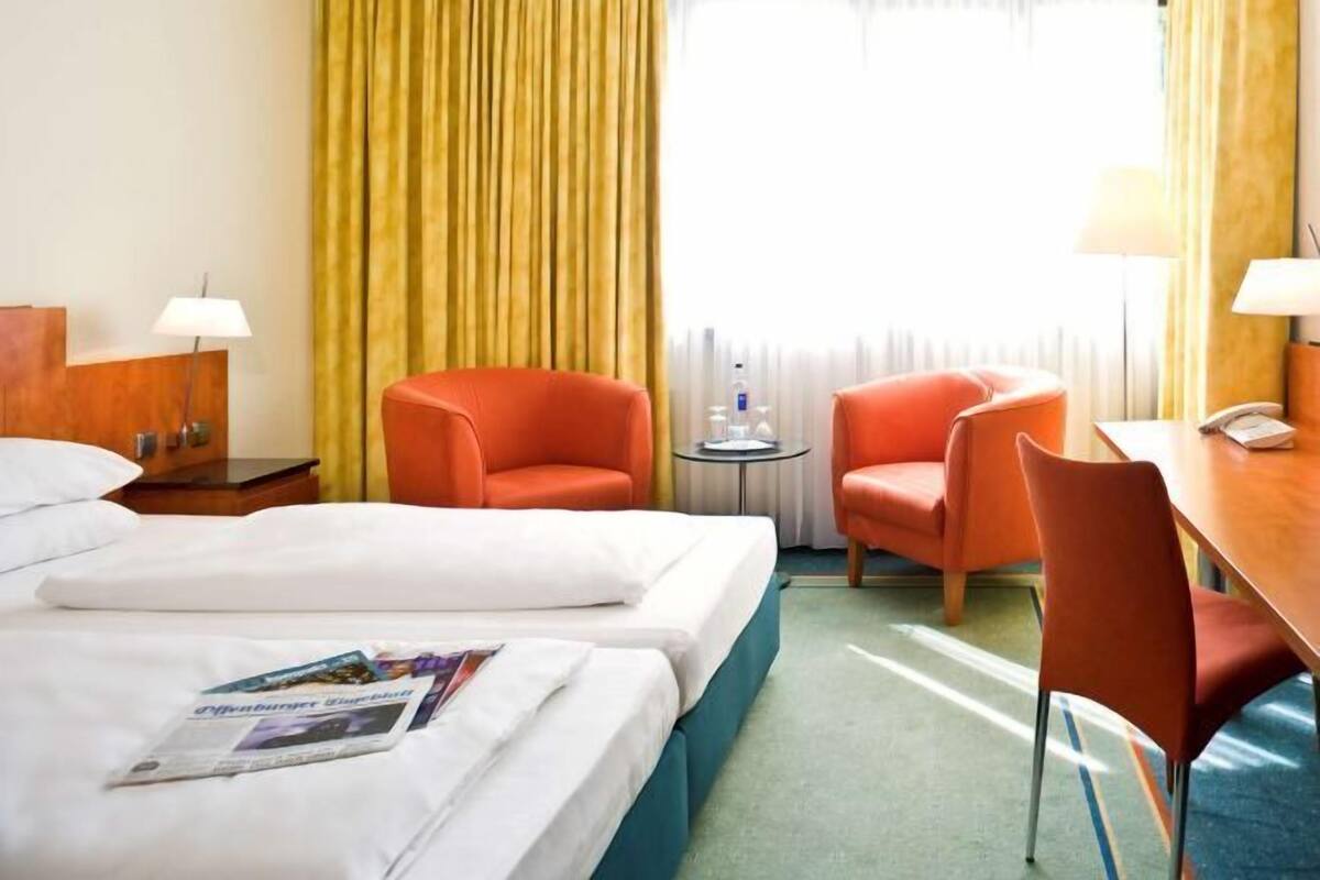 A hotel room with two beds, a desk, two chairs, a lamp, a water bottle, and a window with curtains.