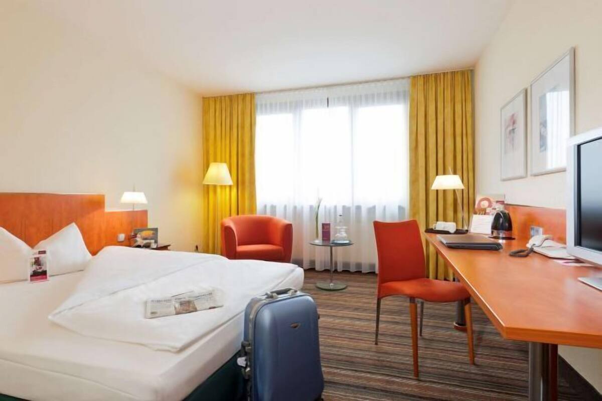 A hotel room with a bed, a desk, two chairs, a television, and a window with curtains.
