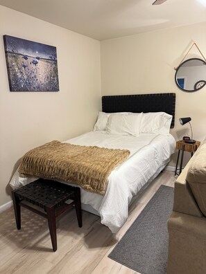A neatly made bed with a white comforter and a brown throw blanket, a bedside table with a lamp, a round mirror, and a painting on the wall.