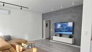En moderne stue med et stort fladskærms-tv monteret på en grå væg, en beige sofa, et lille rundt bord og en trægulv.