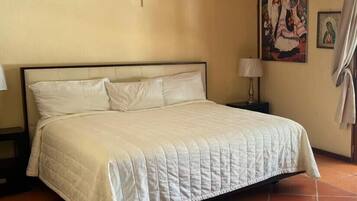 Una cama con ropa de cama blanca, una cruz en la pared y un cuadro colgado.