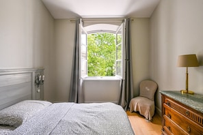 Une chambre à coucher avec un lit, une fenêtre avec des rideaux, une chaise, une commode et une lampe.