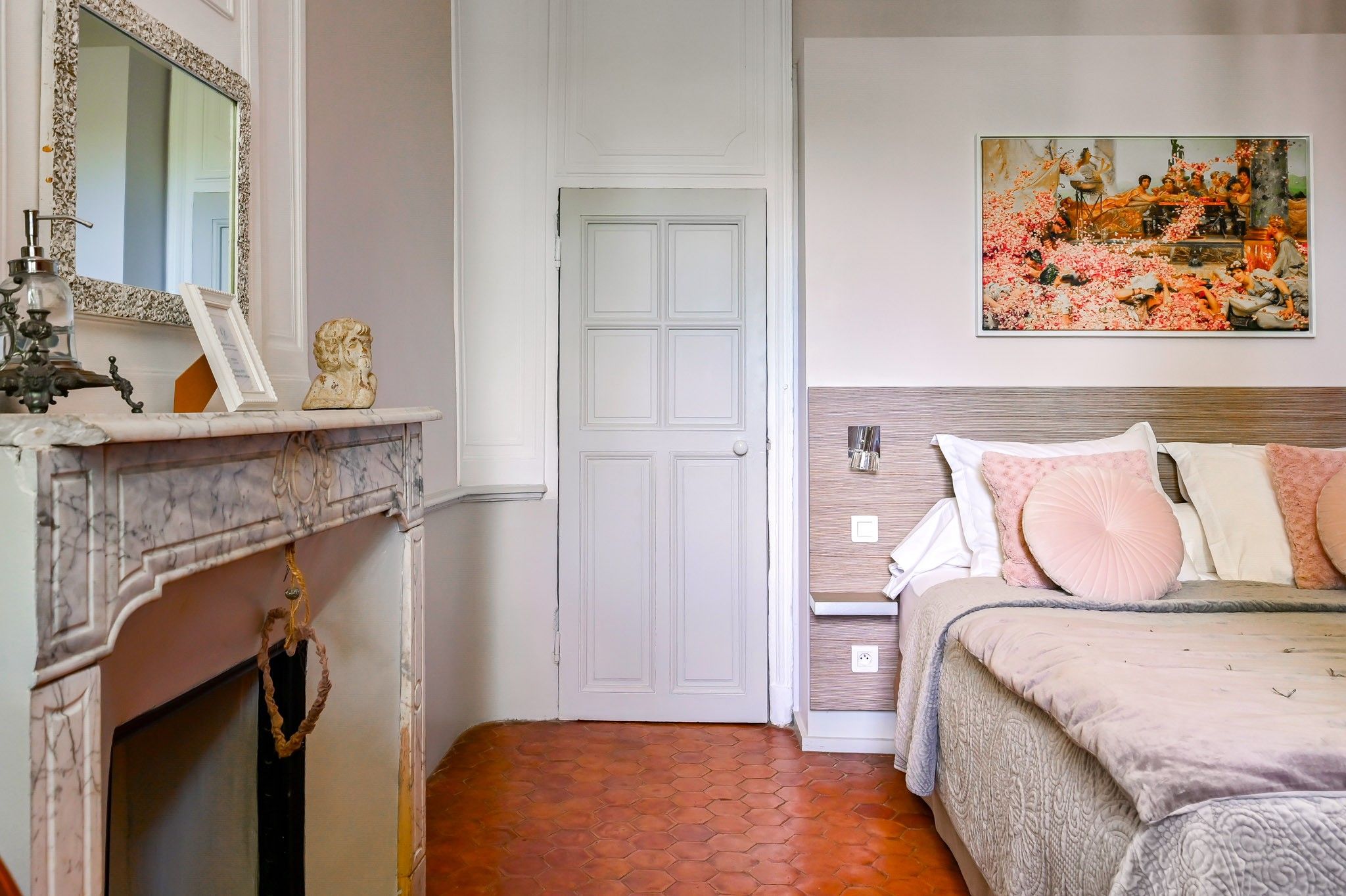 A bedroom with a bed, a nightstand, a painting, a fireplace, and a door.