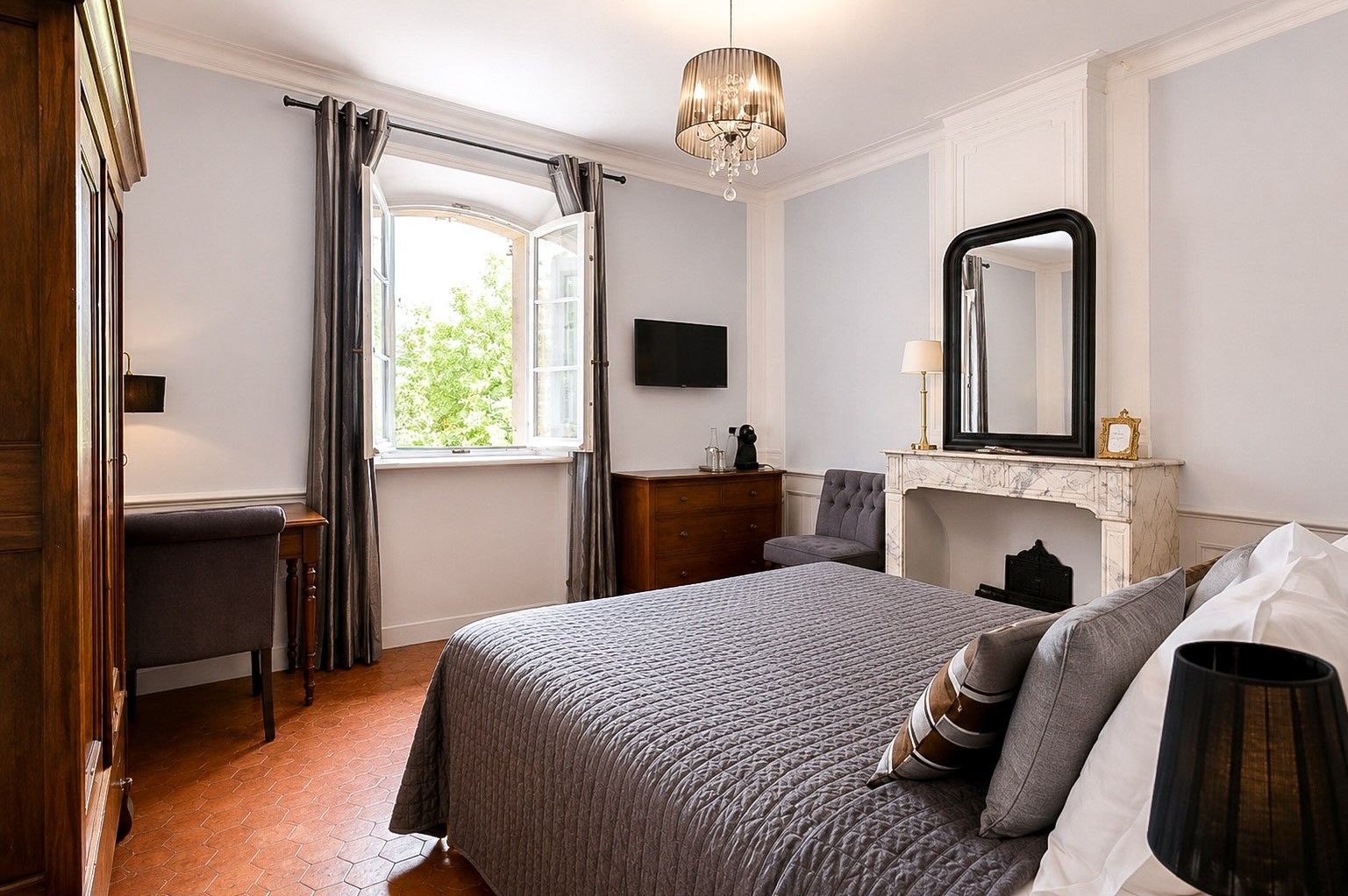 A bedroom with a bed, a dresser, a chair, a fireplace, and a large mirror.