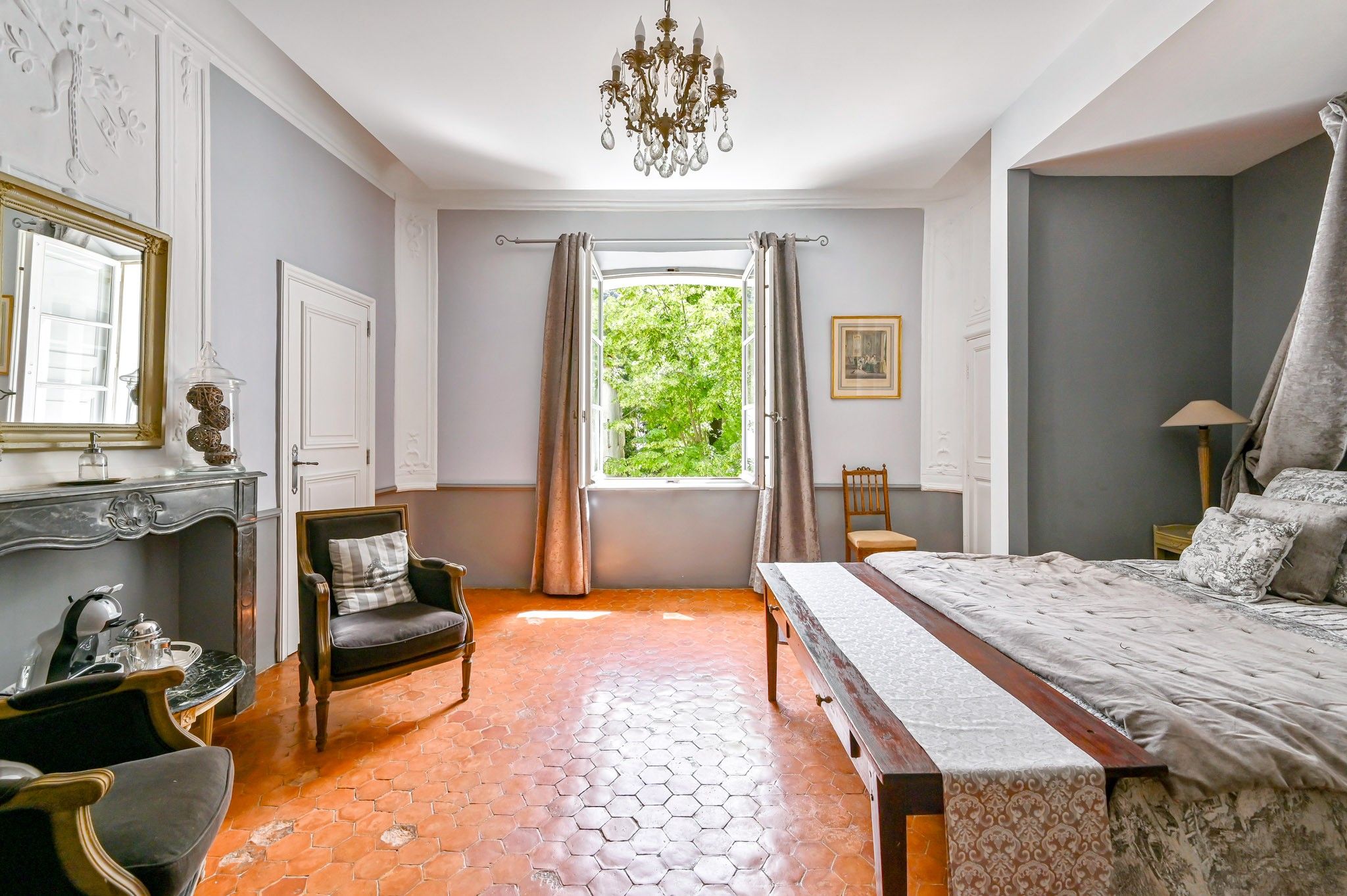 Une chambre à coucher comprenant un lit, une chaise, une table, un lustre, une fenêtre avec des rideaux et une vue sur la verdure à l’extérieur.