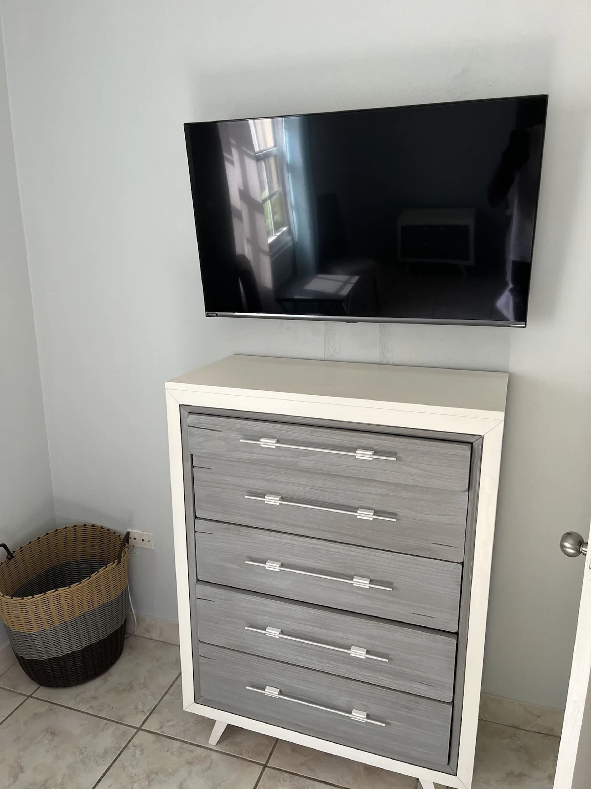 Una habitación con un televisor de pantalla plana montado en la pared, un aparador gris con cajones y una cesta de mimbre en el suelo.