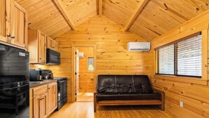 Interior de cabaña con paredes de madera, una zona de cocina con electrodomésticos, un sofá de cuero negro y una ventana con persianas.