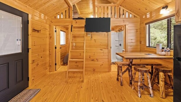 Interior de una cabaña de madera con cocina, una mesa de comedor con taburetes y un televisor montado en la pared.