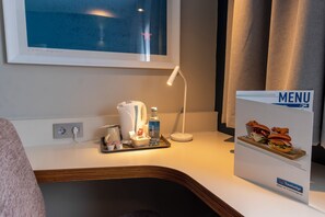 A hotel room with a bedside table featuring a lamp, a water bottle, a coffee maker, and a tray with snacks. A menu sign is visible on the wall. - Travelodge Malaga Aeropuerto (Málaga)