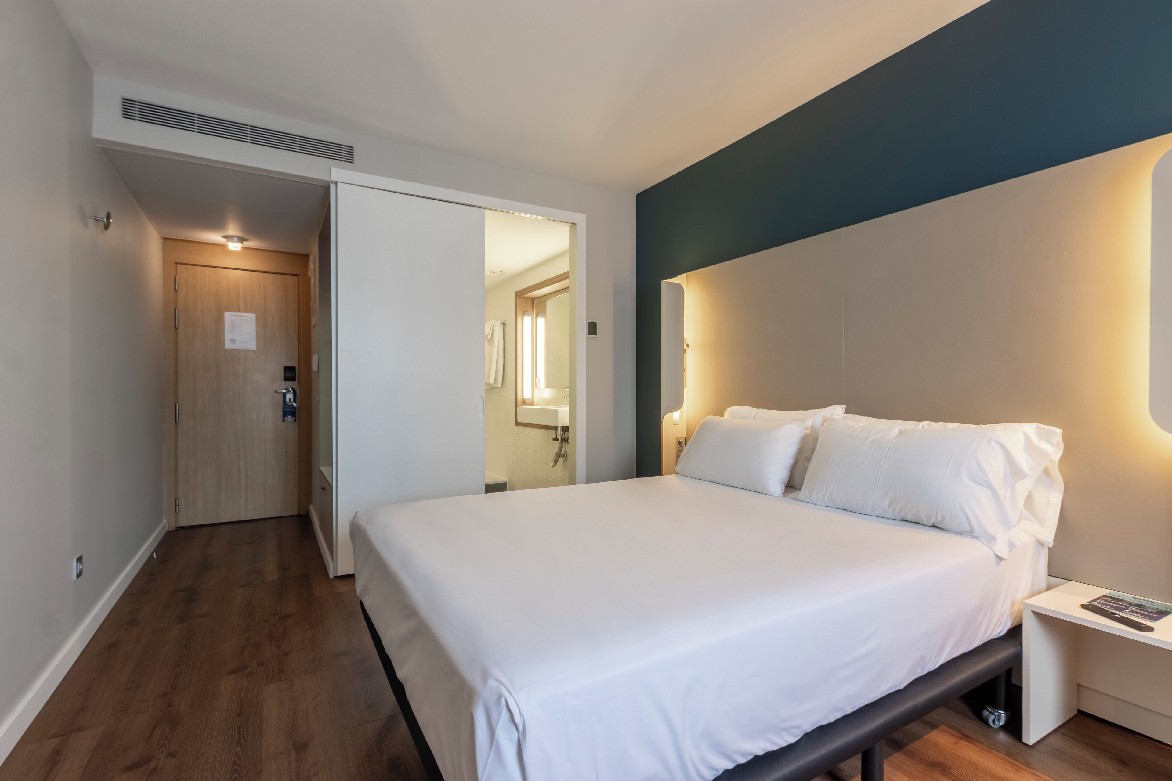 Habitación de hotel con cama, mesita de noche y baño a la vista a través de un acceso abierto.