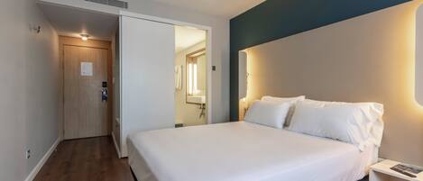 A hotel room with a bed, a bedside table, a nightstand, a wooden door, and a bathroom visible through an open door.