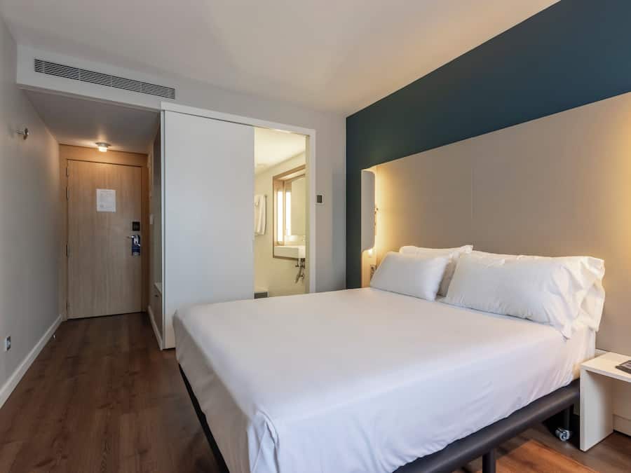 Habitación de hotel con cama, mesita de noche y baño a la vista a través de un acceso abierto.