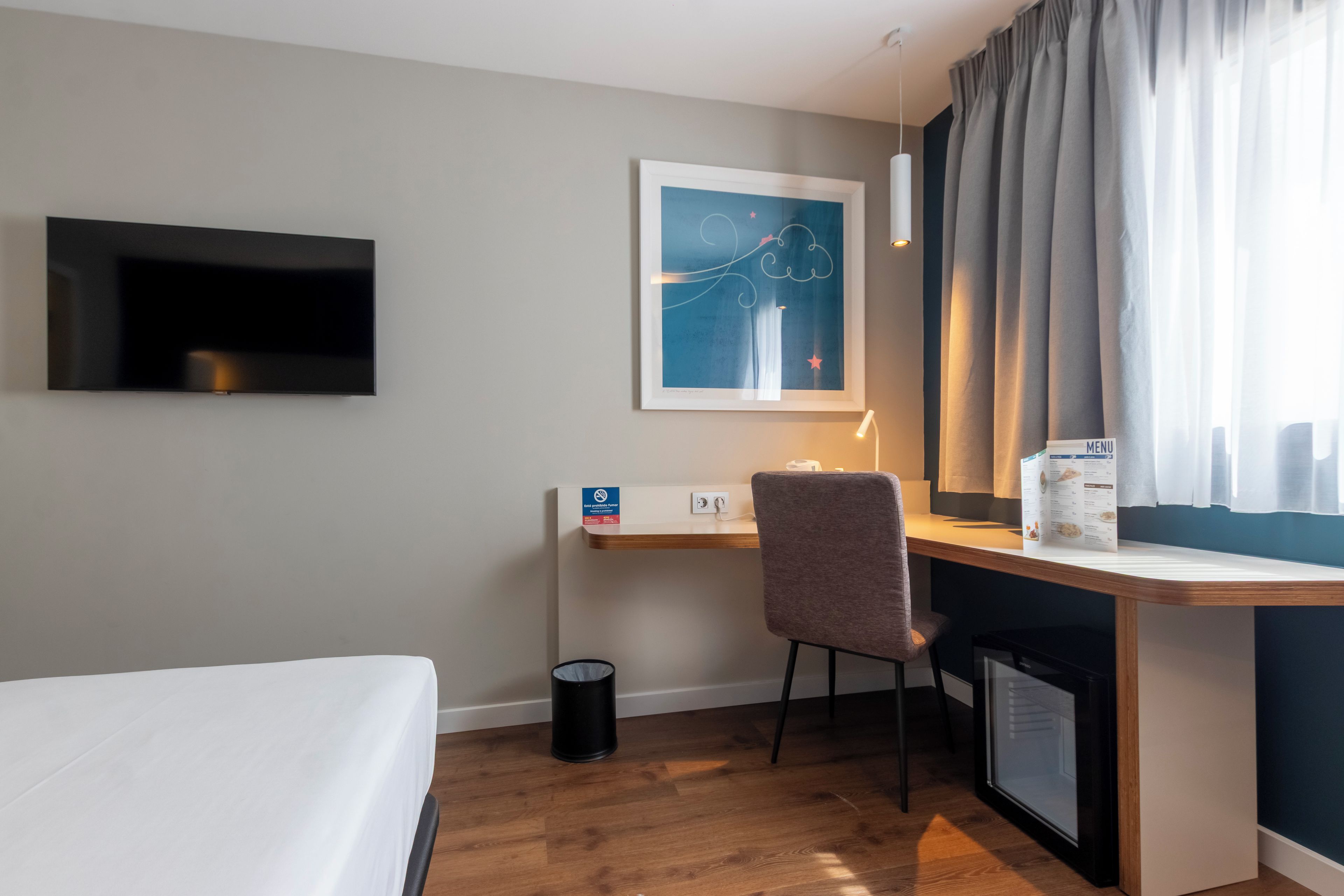 A hotel room with a bed, a desk with a chair, a television mounted on the wall, a framed artwork, a window with curtains, and a small refrigerator.