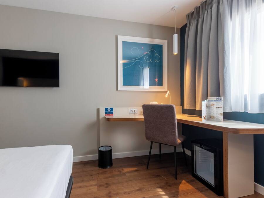 Une chambre d’hôtel comprenant un lit, un bureau avec une chaise, un téléviseur fixé au mur, une œuvre d’art encadrée, une fenêtre avec des rideaux et un petit réfrigérateur.