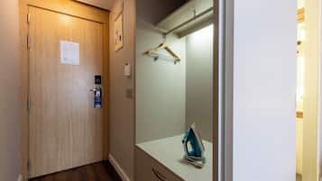 Una habitación de hotel con una puerta de madera, un tocador blanco con espejo, un perchero y un leña azul sobre el tocador.