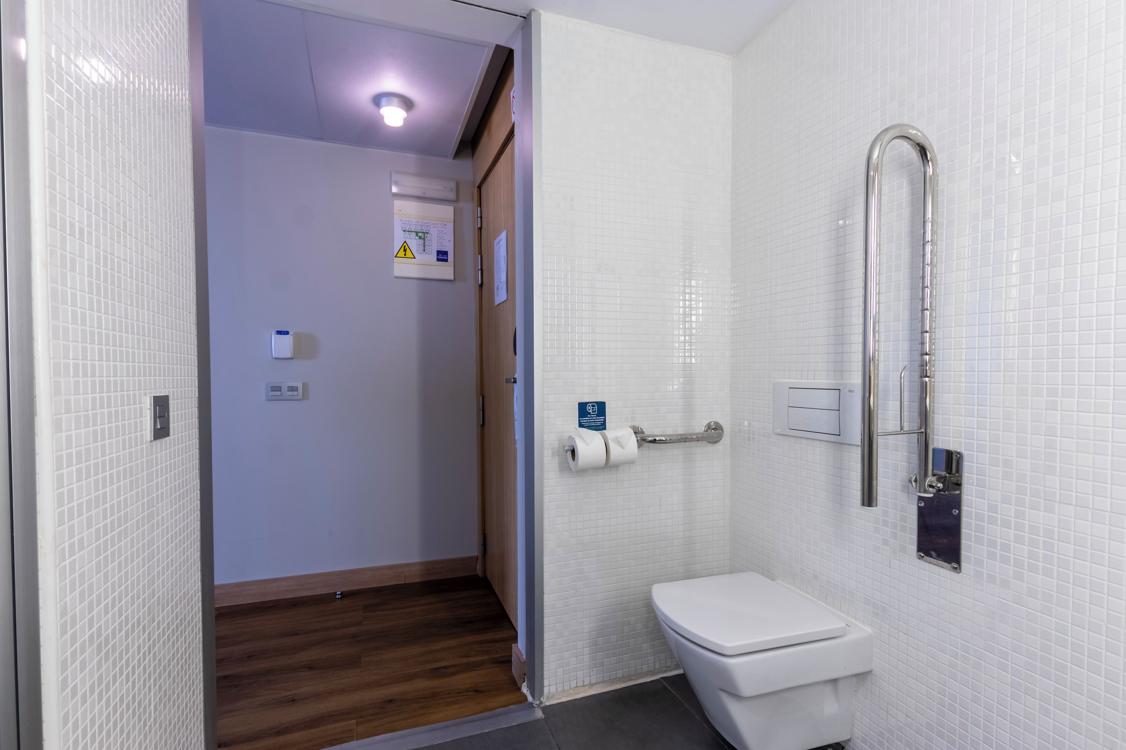 Un baño moderno con paredes de azulejos blancos, suelo de madera, inodoro blanco y ducha con barra de apoyo.