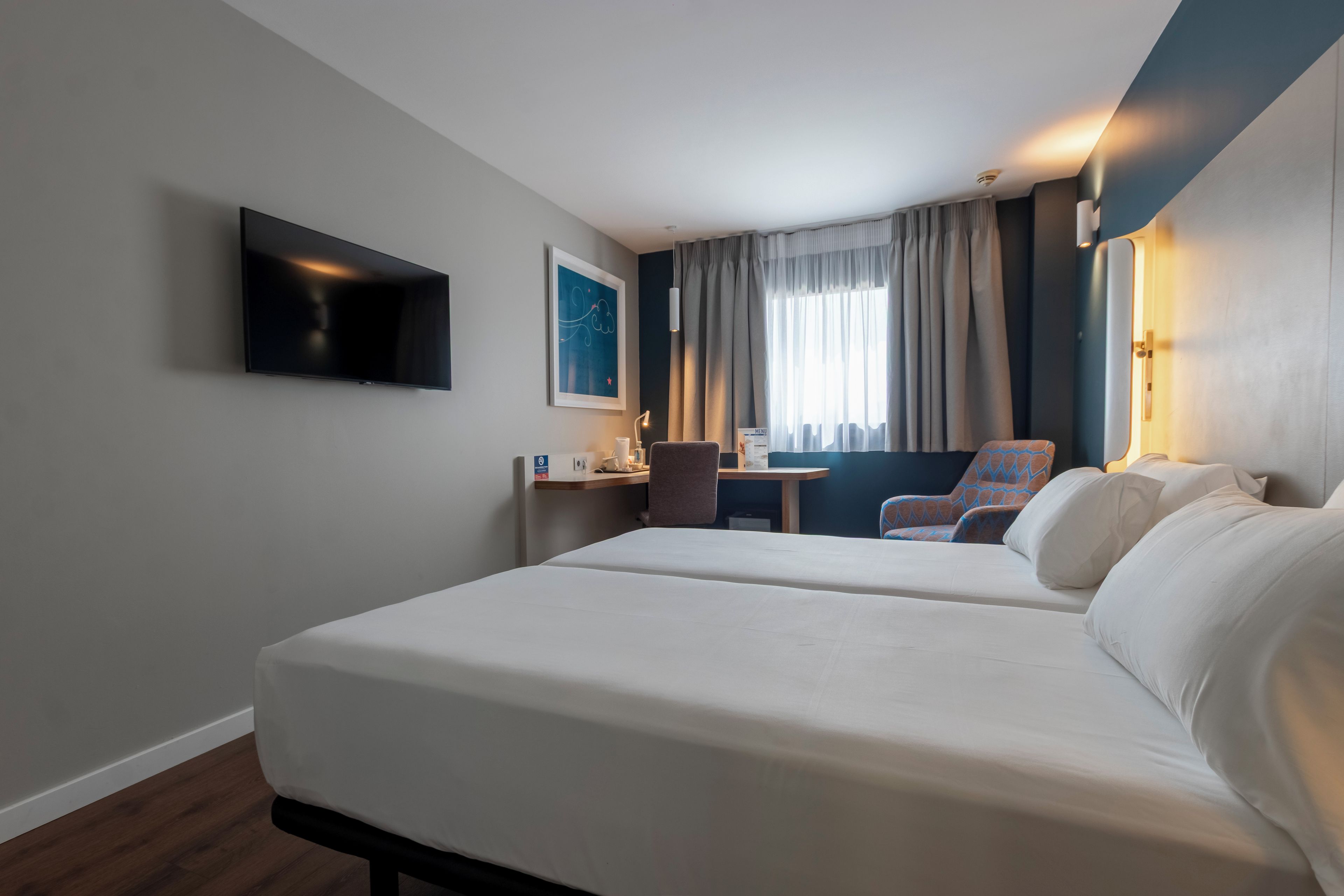 Una habitación de hotel con dos camas, un escritorio, una silla, un televisor y una ventana con cortinas.