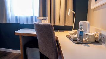 Habitación de hotel con escritorio, una silla, un cesto de basura pequeño, una botella de agua, una cafetera y un cartel con el menú.
