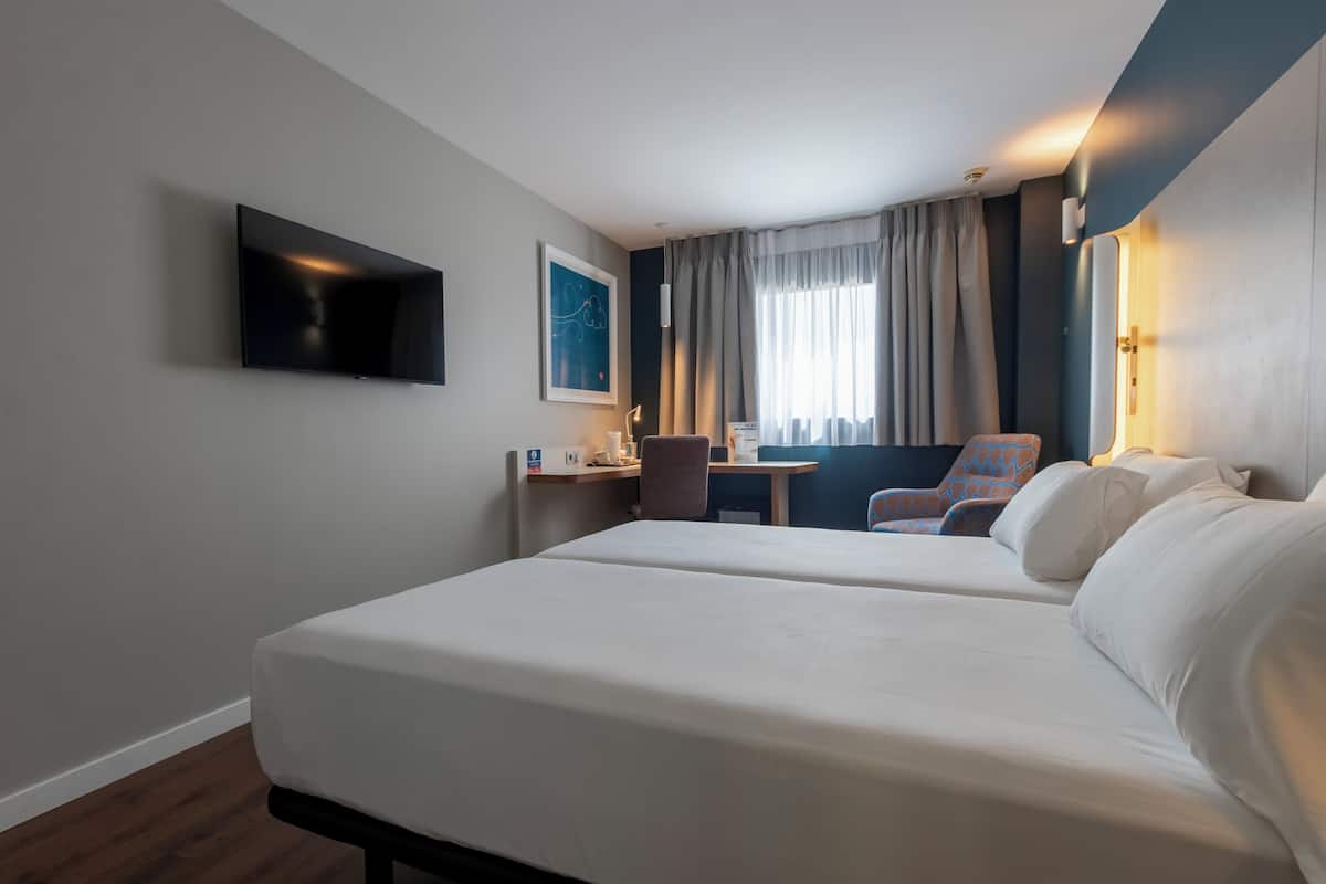 A hotel room with two beds, a desk, a chair, a TV, and a window with curtains.