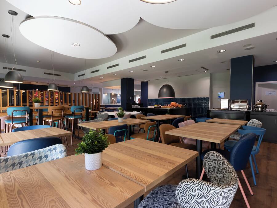 Un restaurant modern amb taules de fusta i cadires blaves, una zona de cuina i una àrea de seients amb un banc estampat.
