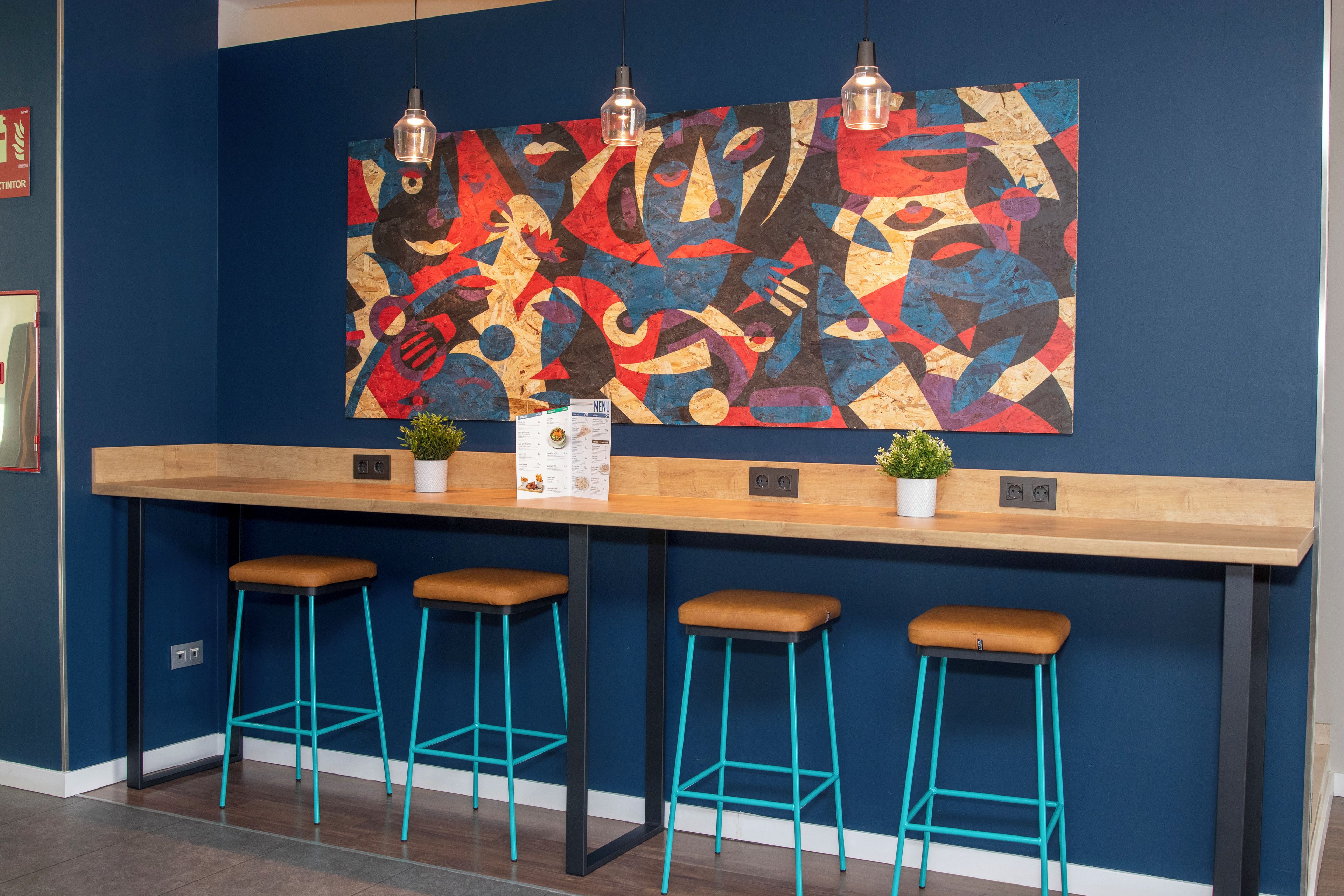 Una zona de bar con mostrador de madera, tres taburetes turquesa y un colorido cuadro abstracto en la pared.