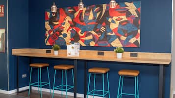 Uma área de bar com balcão de madeira, três banquetas turquesa e uma pintura abstrata colorida na parede.