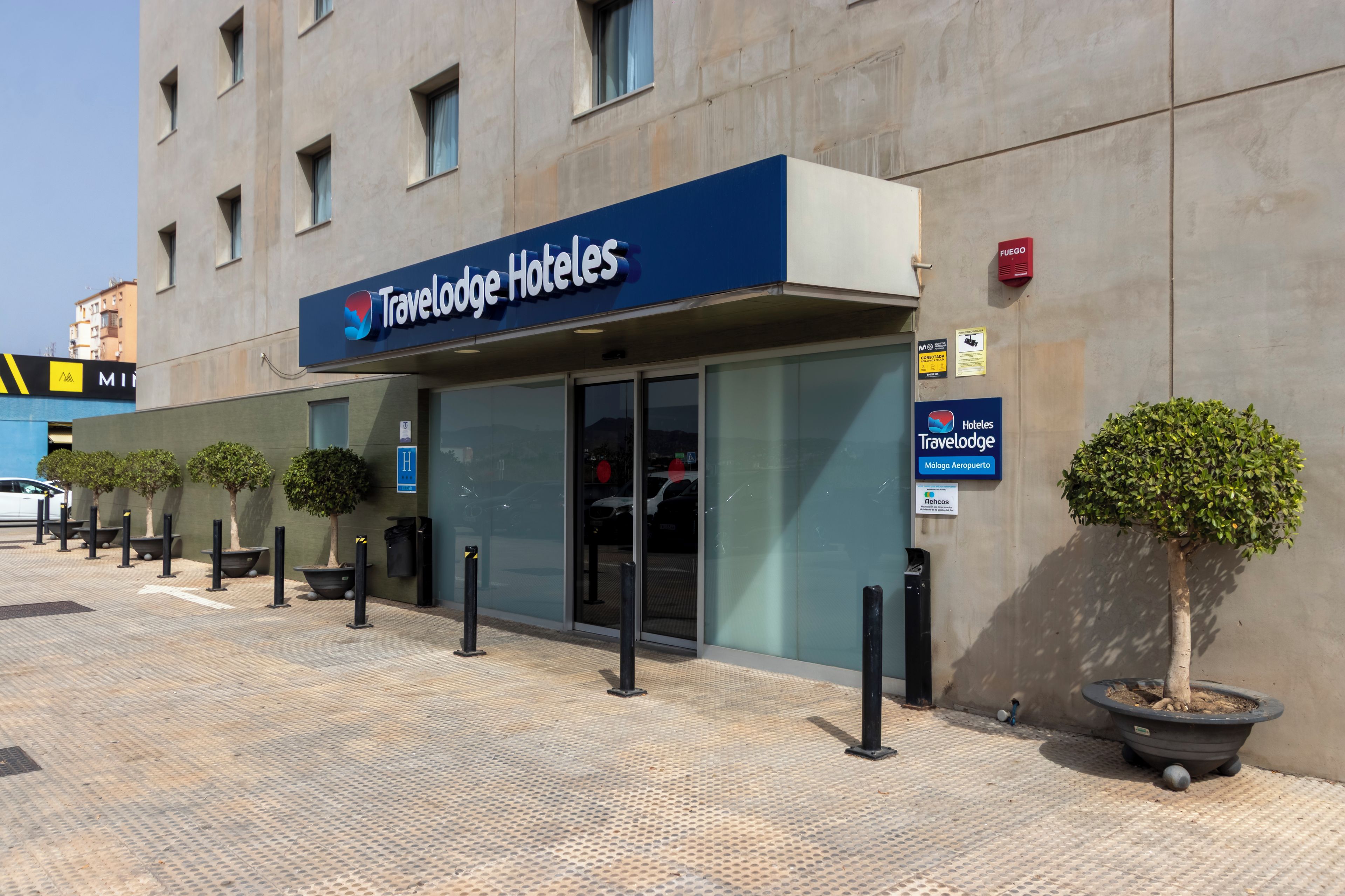 Entrada de un hotel Travelodge con un letrero visible, una zona de estacionamiento y plantas en macetas.
