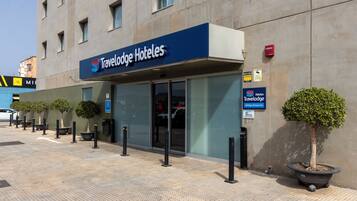 Entrada de un hotel Travelodge con un letrero visible, una zona de estacionamiento y plantas en macetas.