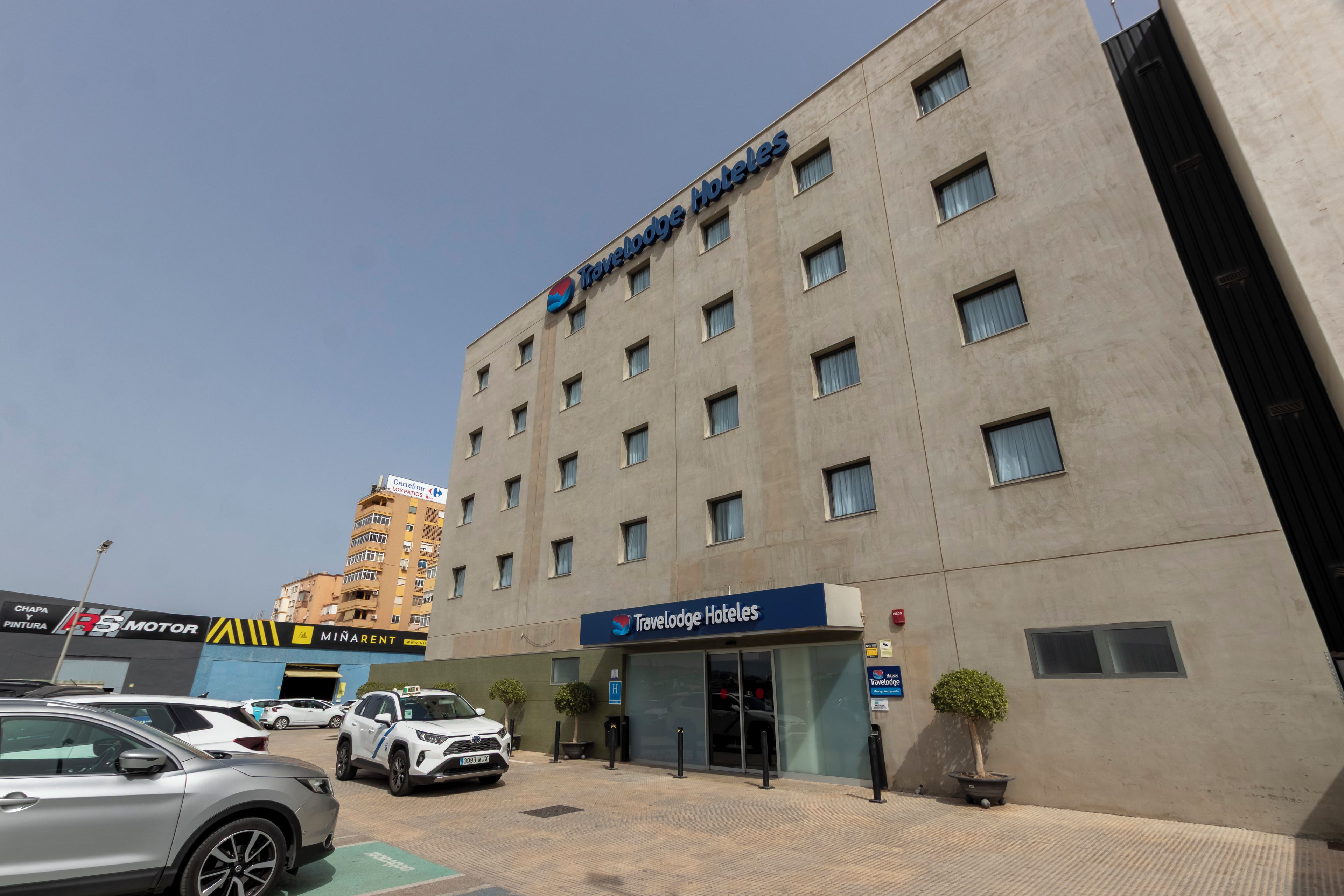 Un hotel Travelodge con fachada moderna, múltiples ventanas y un estacionamiento con automóviles.