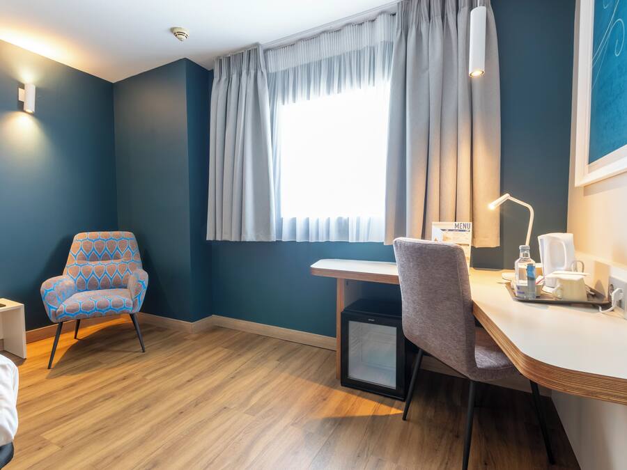 A hotel room with a bed, a desk with a chair, a small fridge, a window with curtains, and a wall-mounted light.