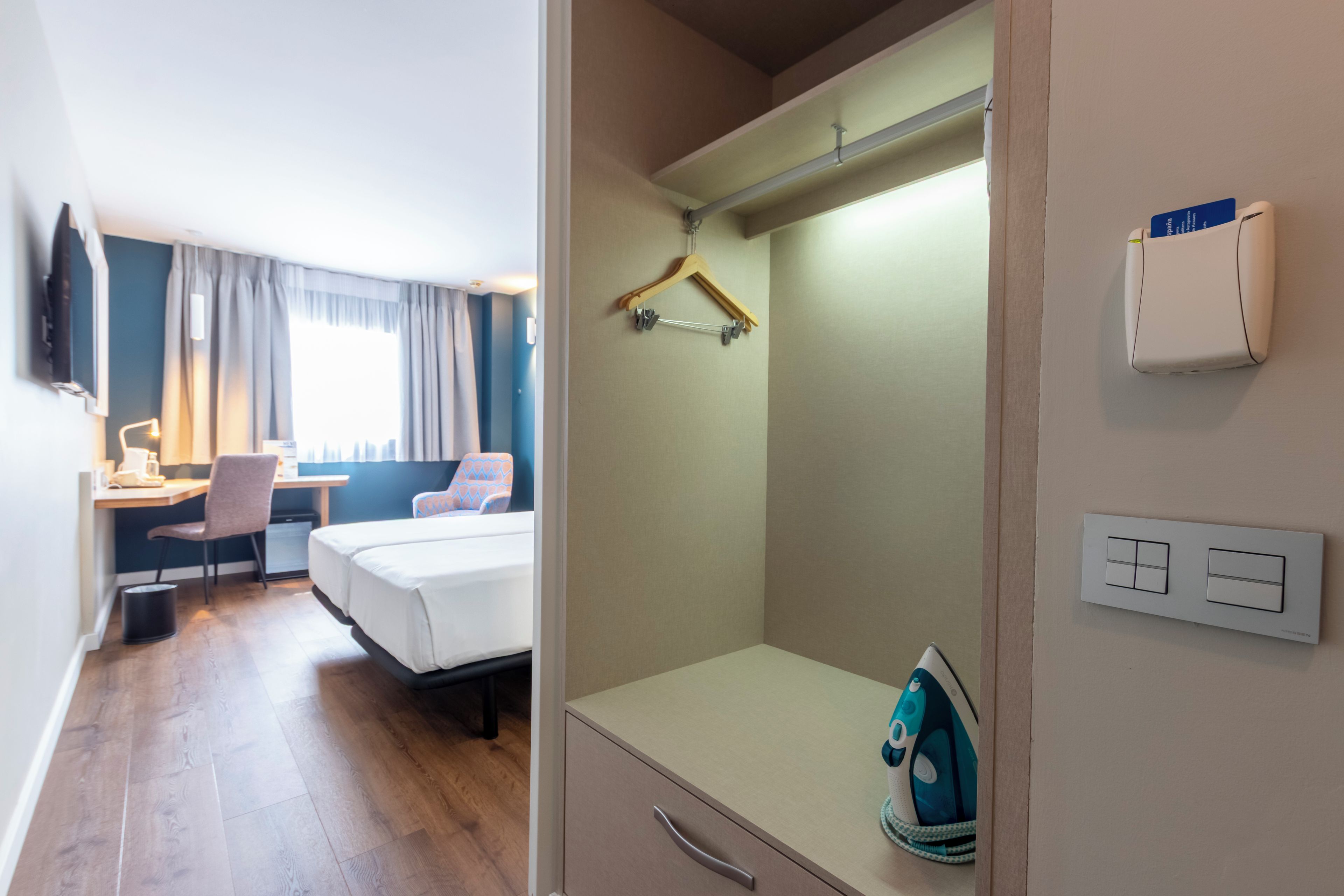 A hotel room with a bed, desk, chair, and a small ironing board with an iron.