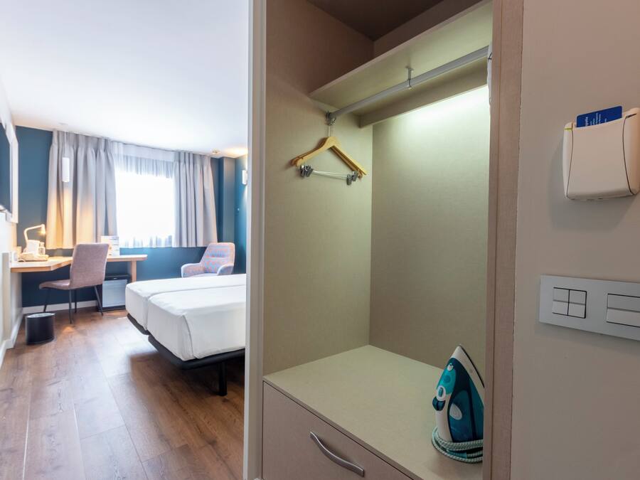 A hotel room with a bed, desk, chair, and a small ironing board with an iron.