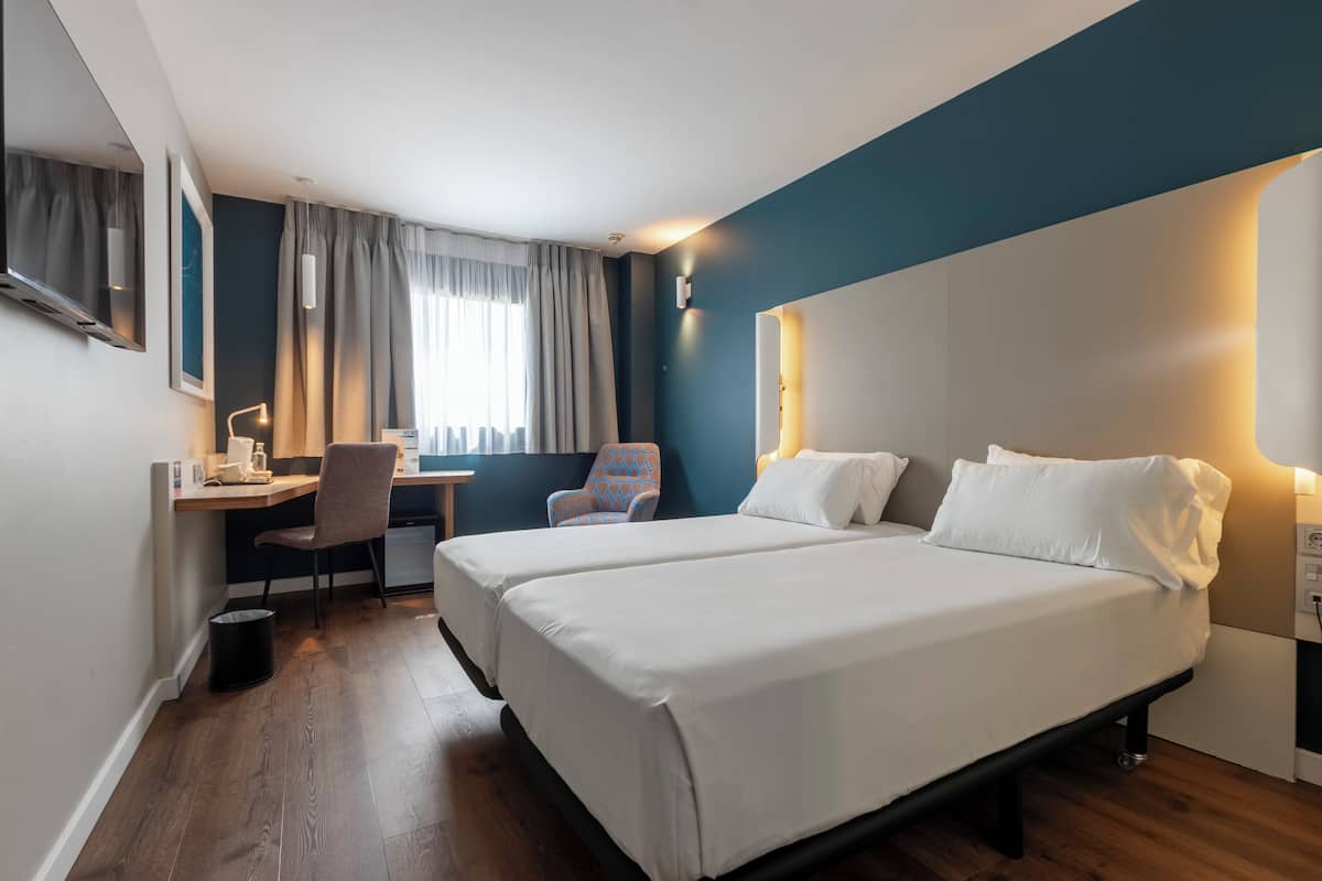 A hotel room with two beds, a desk with a chair, a TV, and a window with curtains.