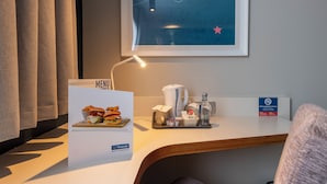 Une chambre d’hôtel avec un bureau blanc, un menu encadré, une machine à café, une bouteille et un panneau indiquant qu’il est interdit de fumer.