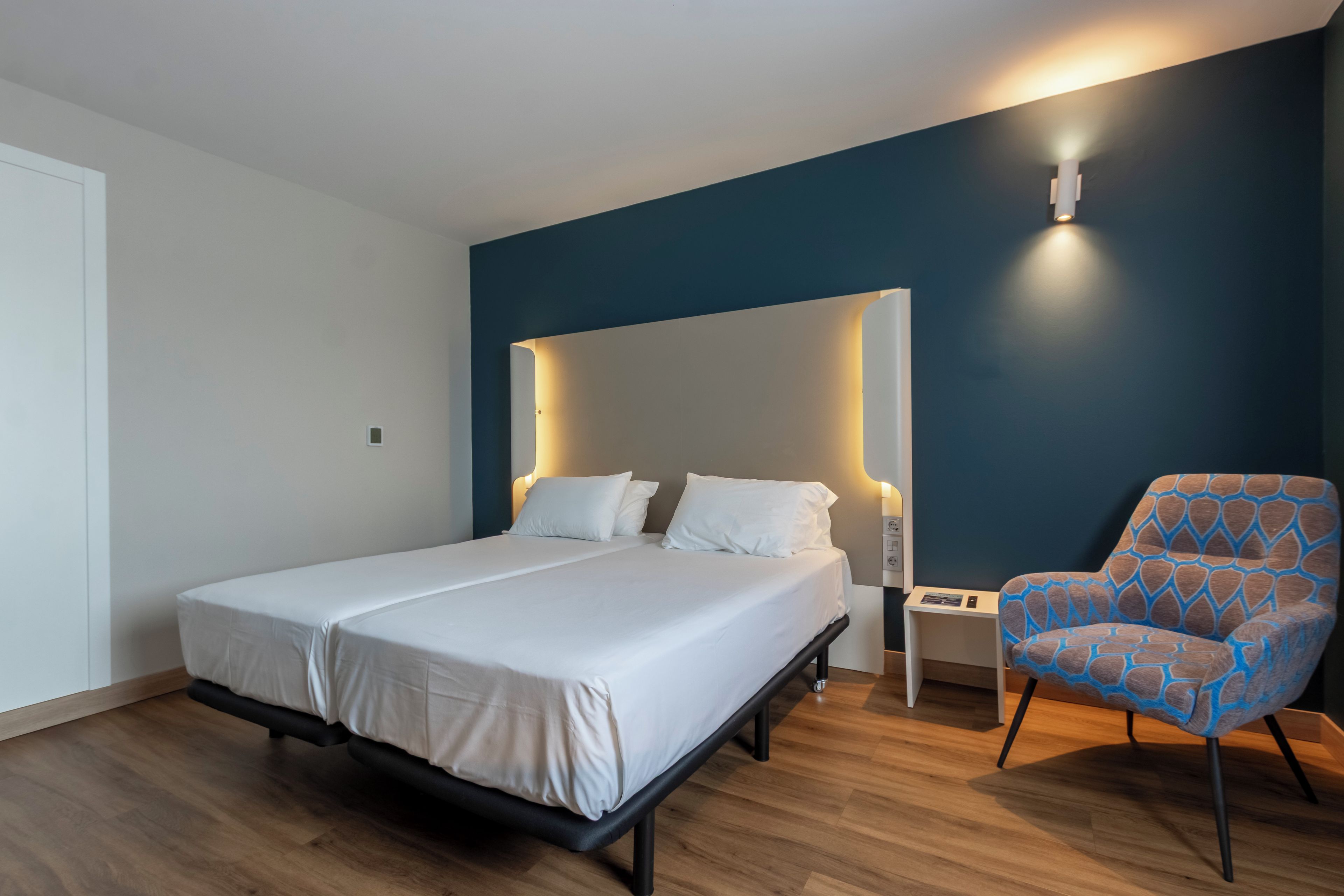 Un dormitorio moderno con una cama grande, una mesita de noche, una silla y una lámpara fijada a la pared.