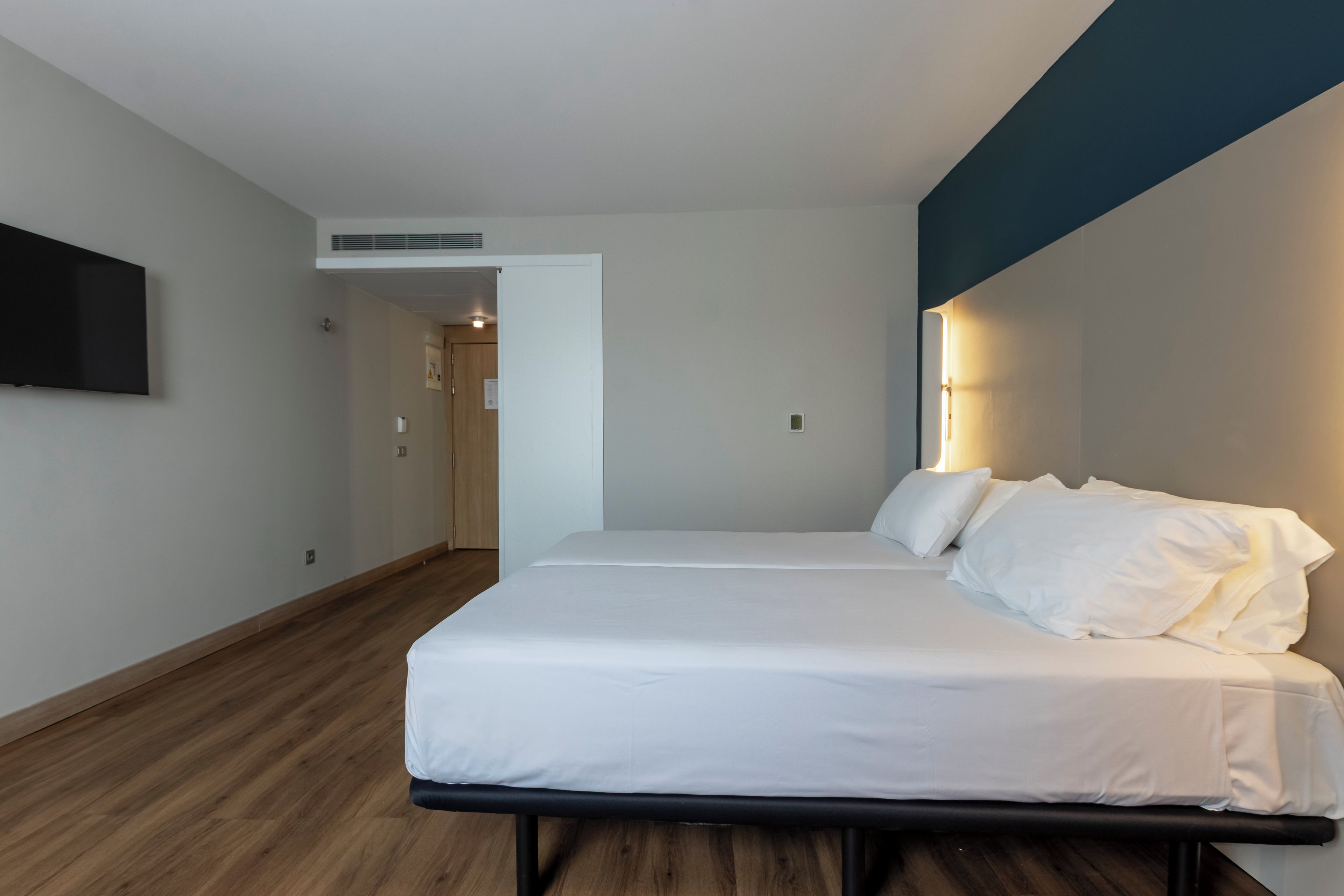 A modern hotel room with a large bed, a flat-screen TV mounted on the wall, and a wooden floor.