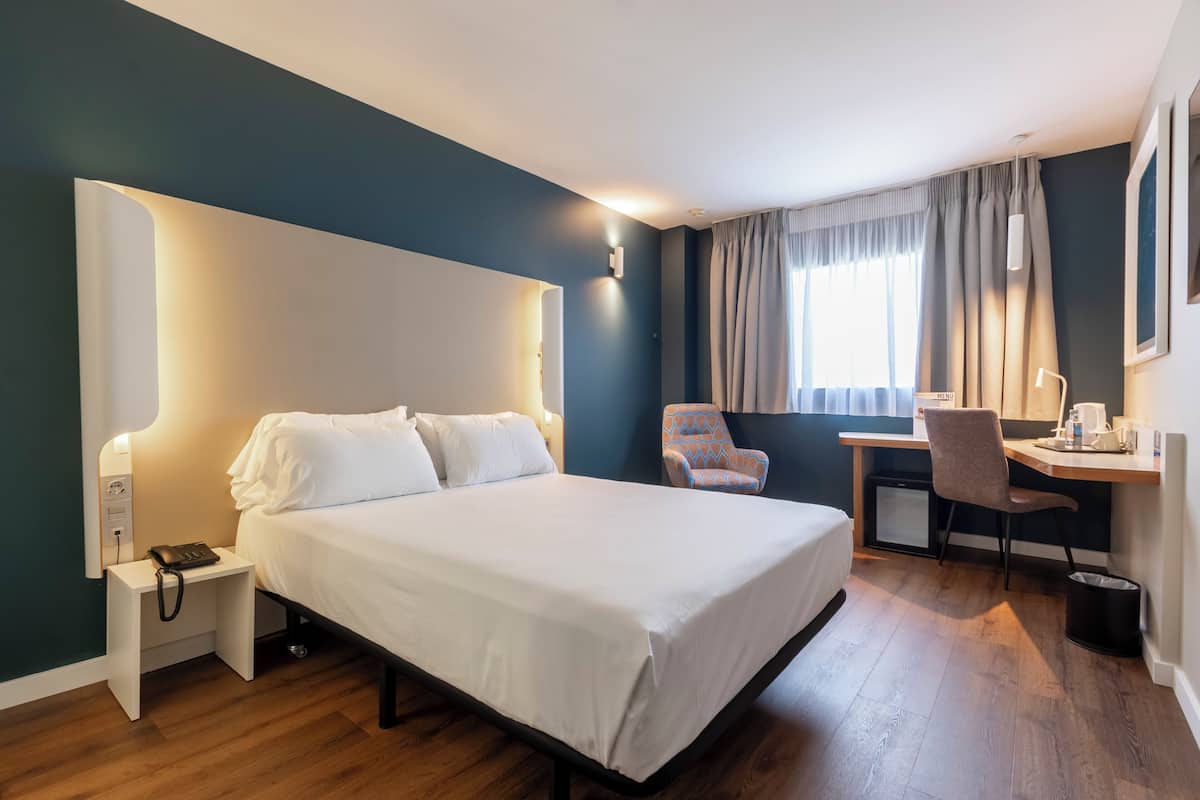 A hotel room with a bed, bedside table, desk, chair, and a small kitchenette.