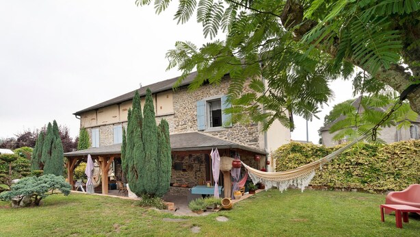 Uma casa de pedra com alpendre, uma rede e um jardim bem cuidado.