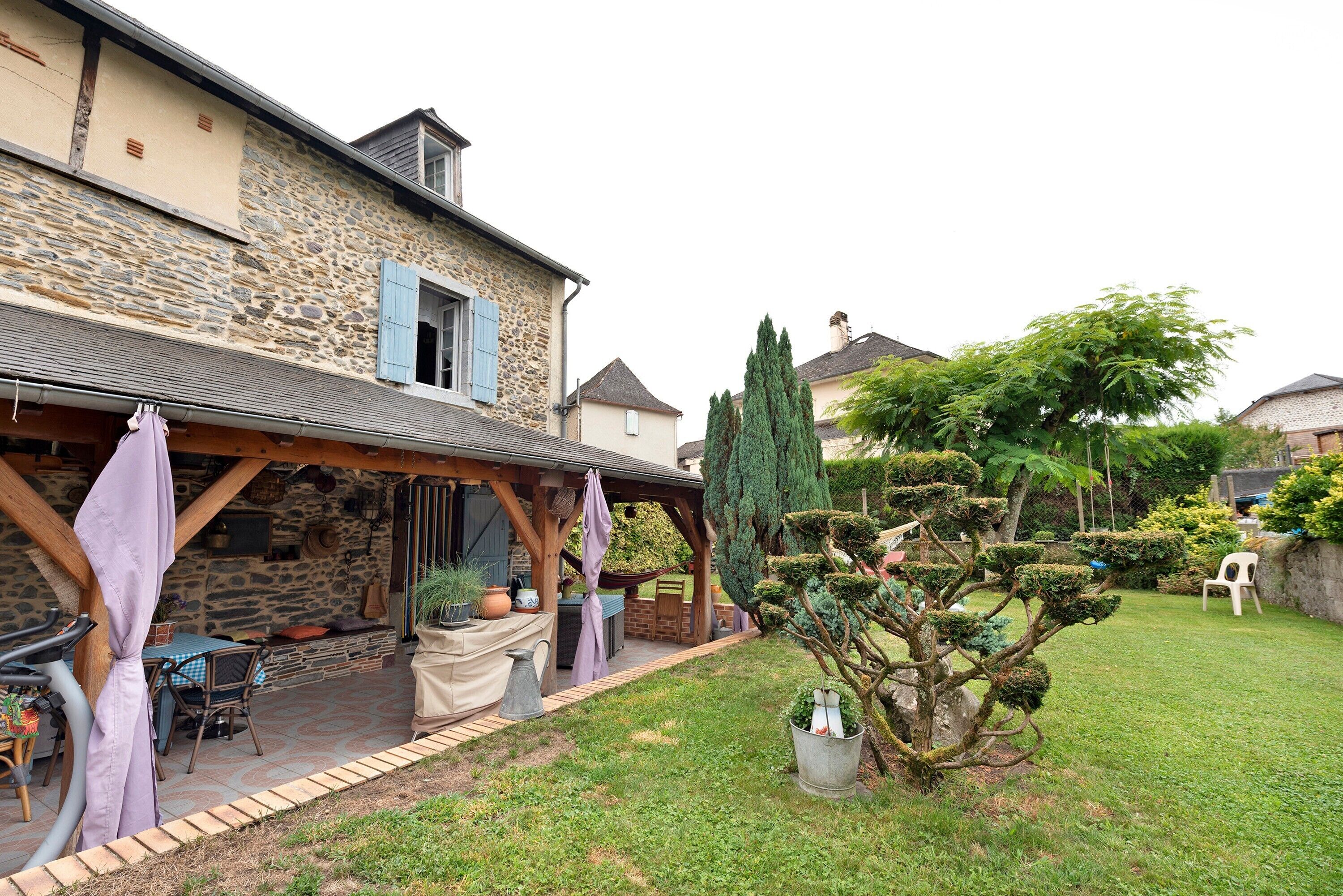 A stone house with a covered patio, outdoor seating, and a well-maintained garden with various plants and trees.